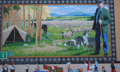 Vale_Basque_Sheepherder_mural A colorful mural of an older gentlemen with working dogs at his side and a herd of sheep behind him. The man is a Basque sheepherder. Small figures dressed in traditional Basque clothing frame the bottom of the mural.